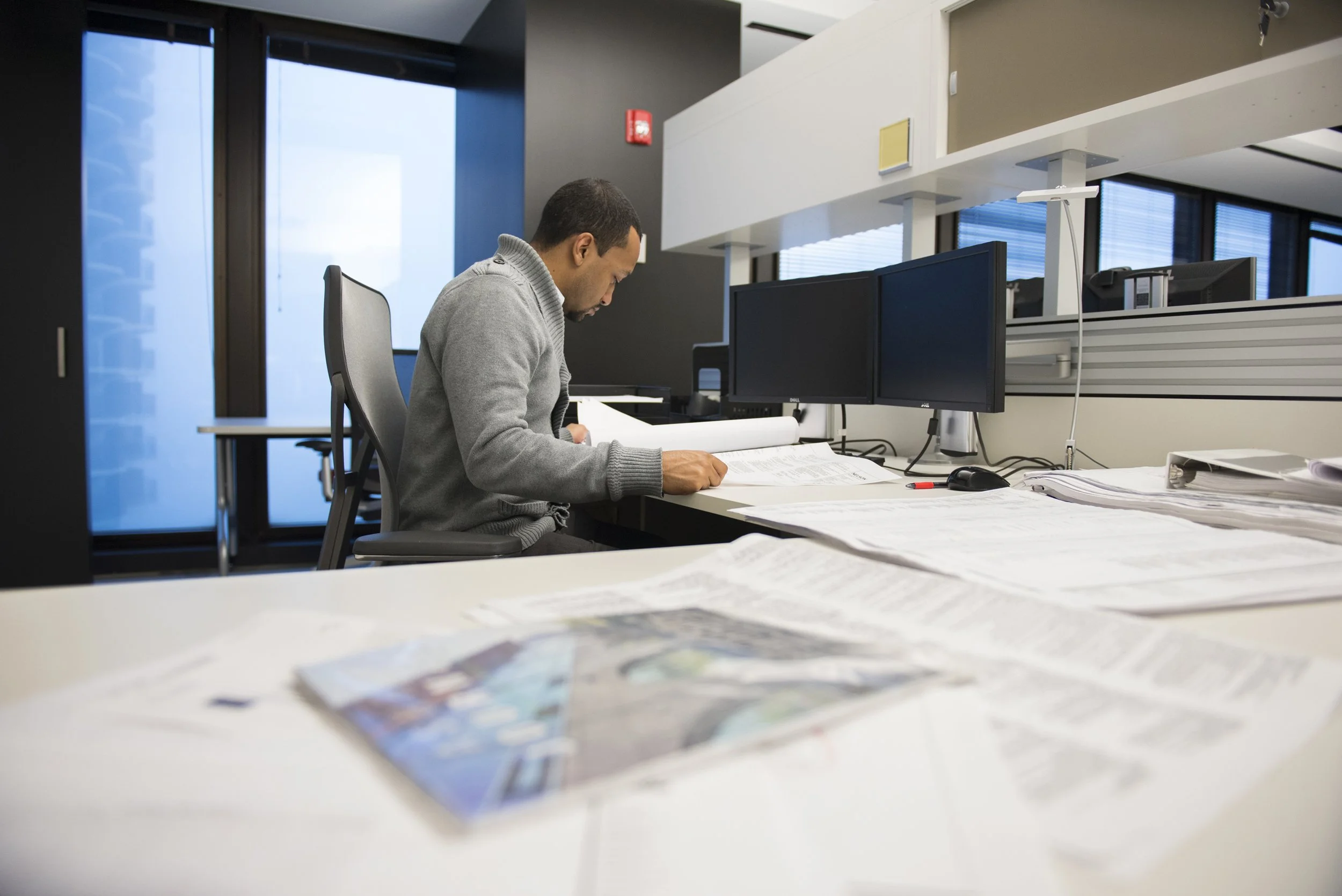 A man sitting at a desk in an office, studying documents with multiple computer monitors and a stack of papers.