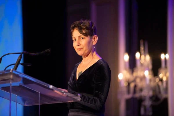 A woman with short dark hair standing behind a clear podium at a formal event, with candles and chandeliers in the background.