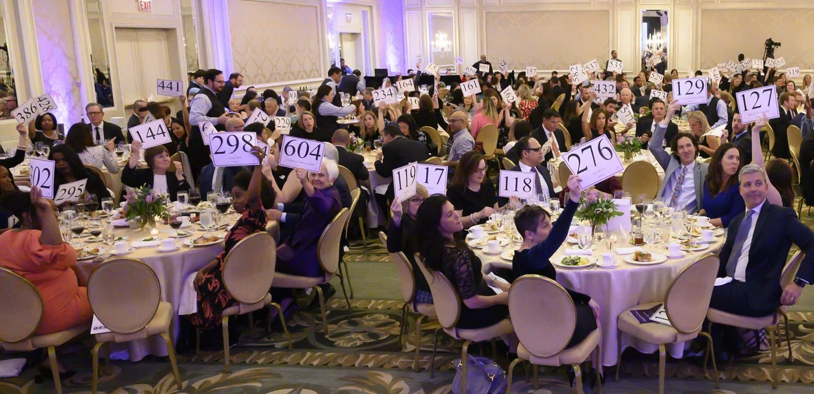 People seated at round tables in a banquet hall participating in an auction, holding up numbered paddles.
