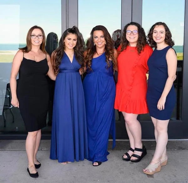 Five women standing together in front of a large window, smiling and dressed in colorful dresses.