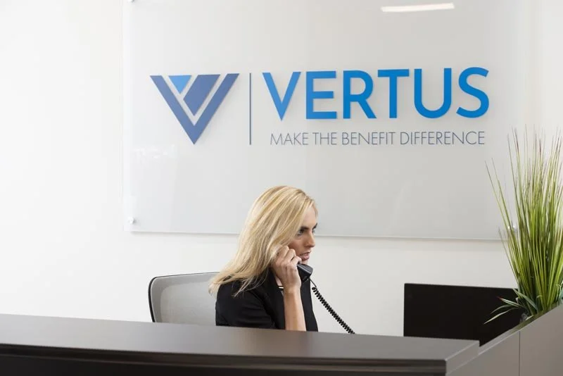 A woman with blonde hair sitting at a reception desk, speaking on a landline phone, in front of a sign that says 'VERTUS' and includes the tagline 'MAKE THE BENEFIT DIFFERENCE' with a logo.
