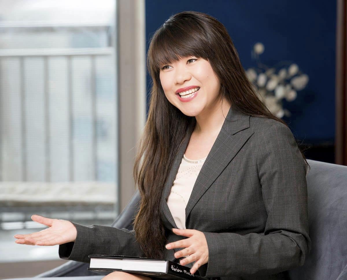 A woman with long brown hair, wearing a gray blazer and white shirt, smiling and gesturing with her hands while sitting indoors.