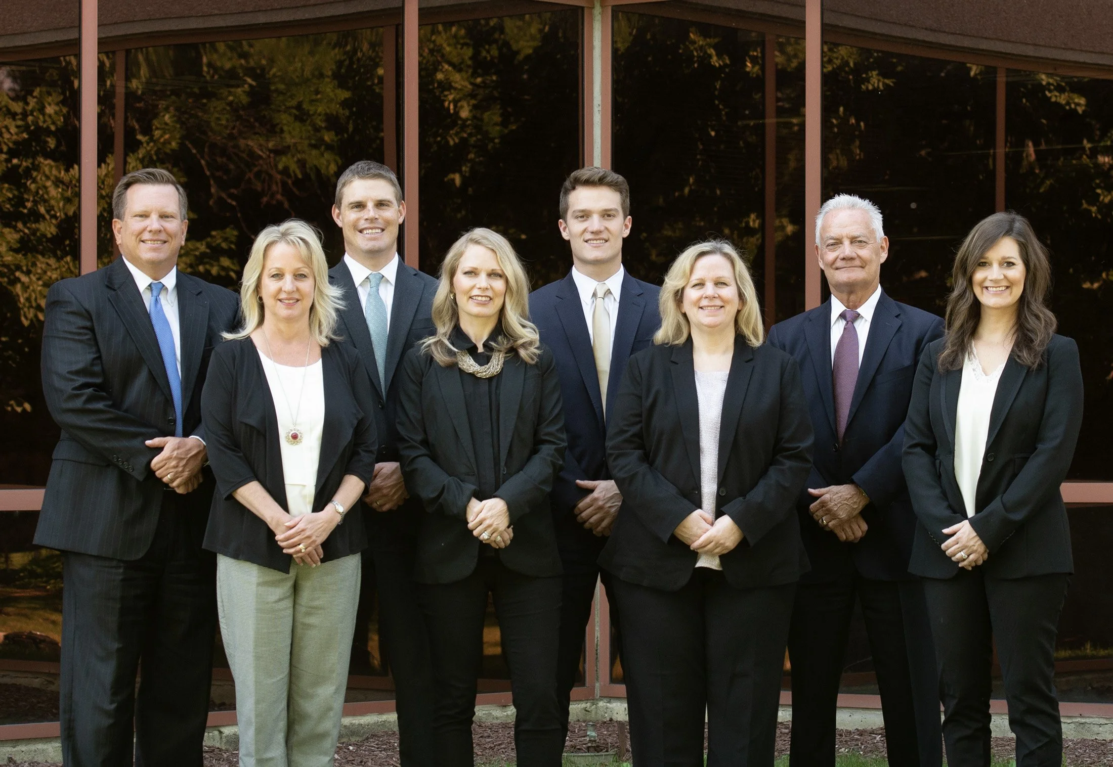 A group photo of professional consultants standing outdoors in front of a glass building with trees in the background.