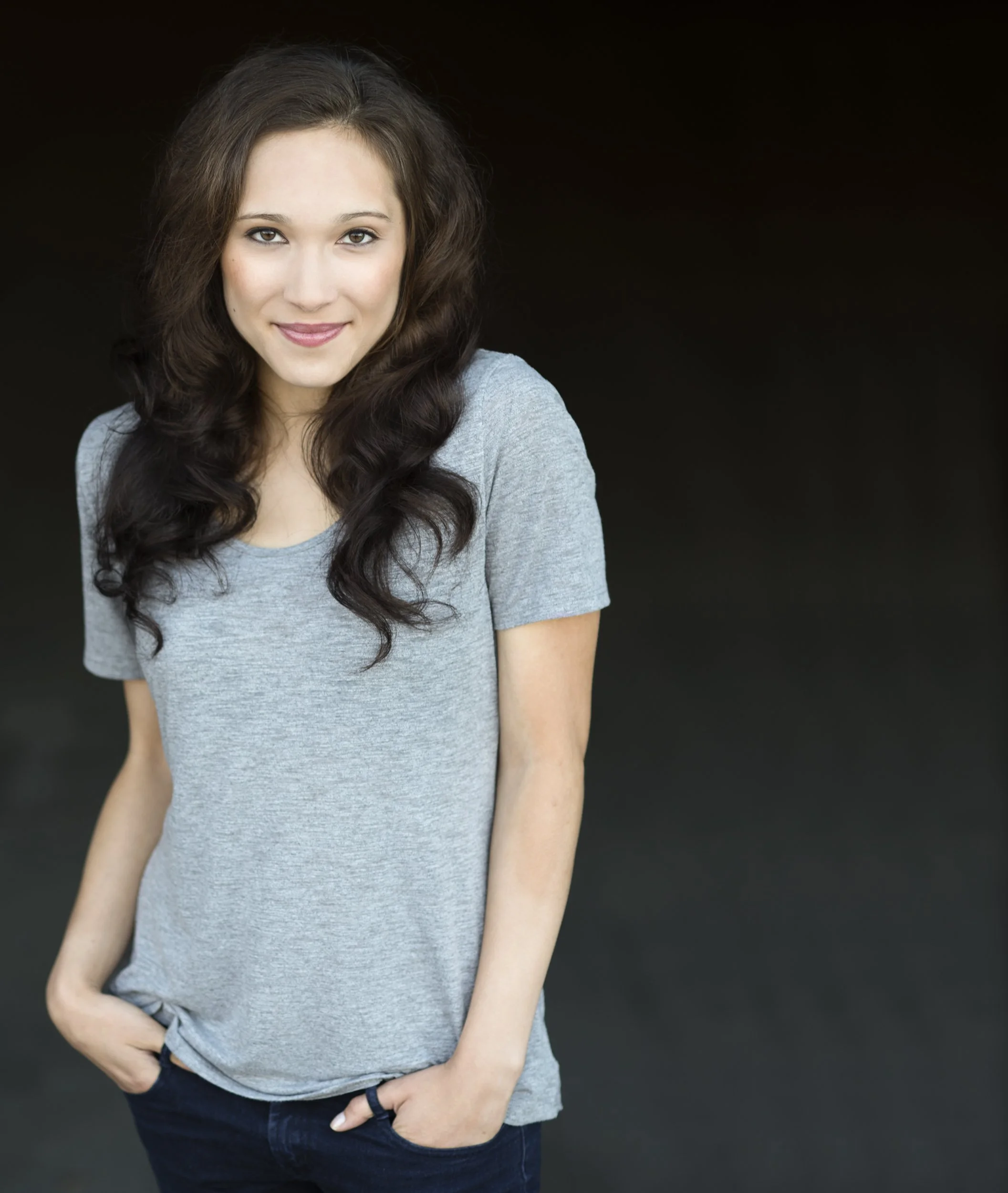 Young woman with long, curly brown hair, wearing a casual gray T-shirt and dark jeans, smiling and posing against a dark background.