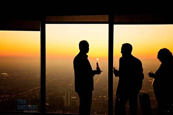 Silhouettes of three people holding drinks, standing near large windows with a city view at sunset.