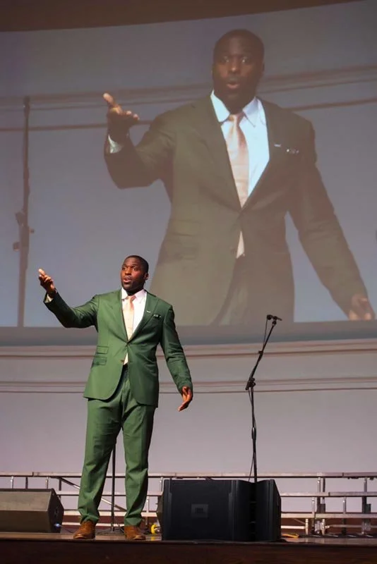 A man in a green suit standing on a stage, speaking or presenting, with a large screen behind him projecting his image during an event at Navy Pier captured by a Chicago photographer