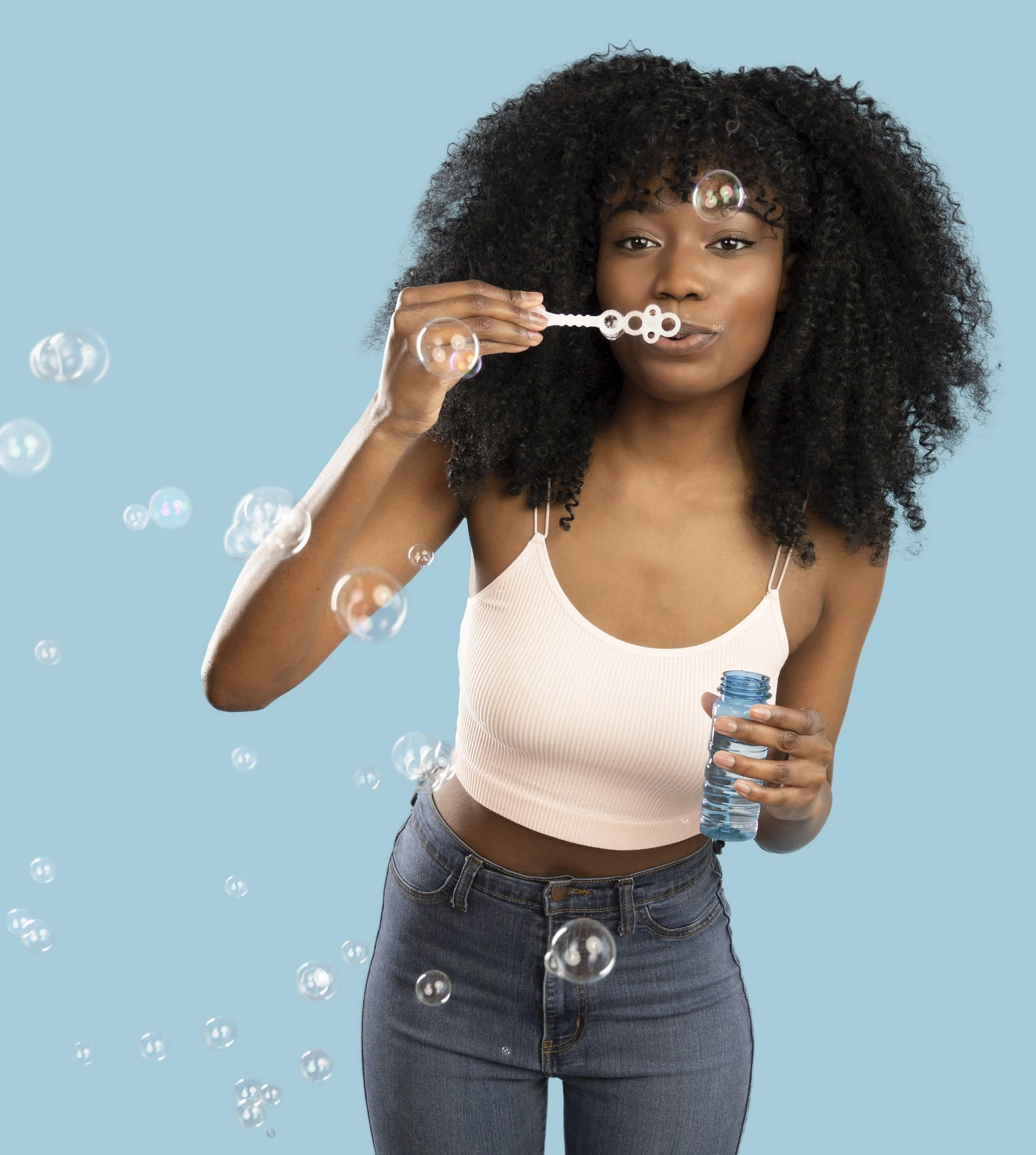 A woman with curly black hair blowing bubbles with a bubble wand, holding a bottle of bubble solution, against a blue background.