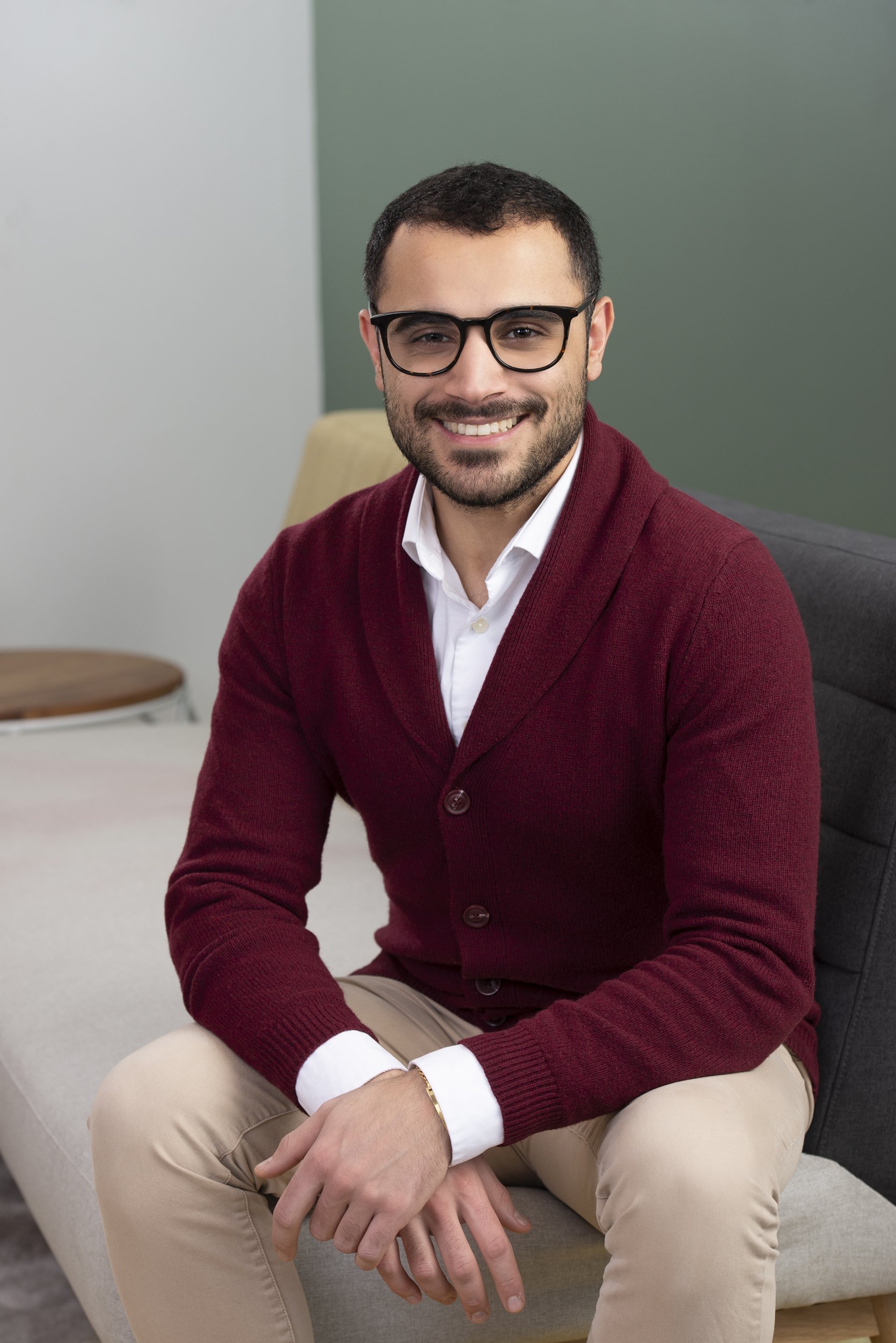 Portrait of a smiling man with dark hair and glasses, wearing a white shirt, burgundy cardigan, and beige pants, sitting on a beige chair in a room with a green wall.