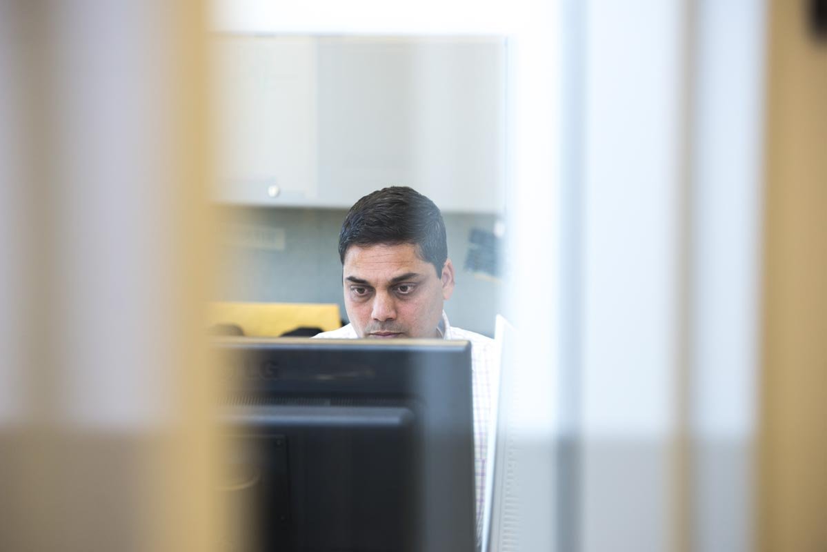 A man working at a computer seen through a small opening between two vertical surfaces.