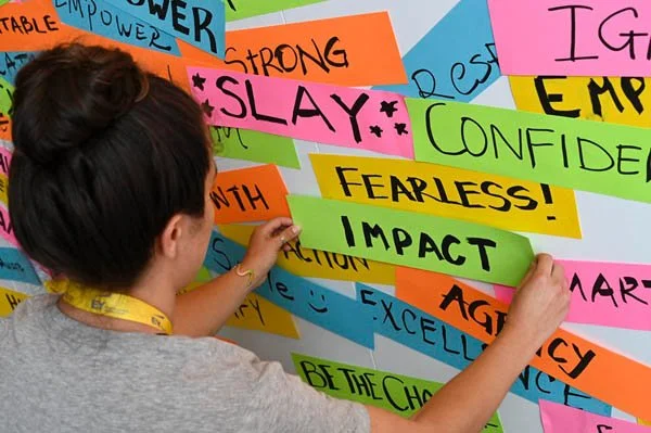 A person with dark hair tied back, wearing a gray shirt and yellow lanyard, placing colorful words related to empowerment and positivity on a vibrant wall display. The words include 'SLAY,' 'FEARLESS,' 'IMPACT,' 'CONFIDENCE,' and others in bright col