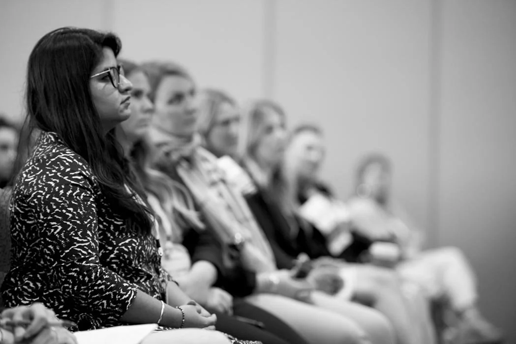 A group of women sitting in a conference room, attentively listening to a presentation, with a woman in the foreground wearing glasses and a patterned blouse.