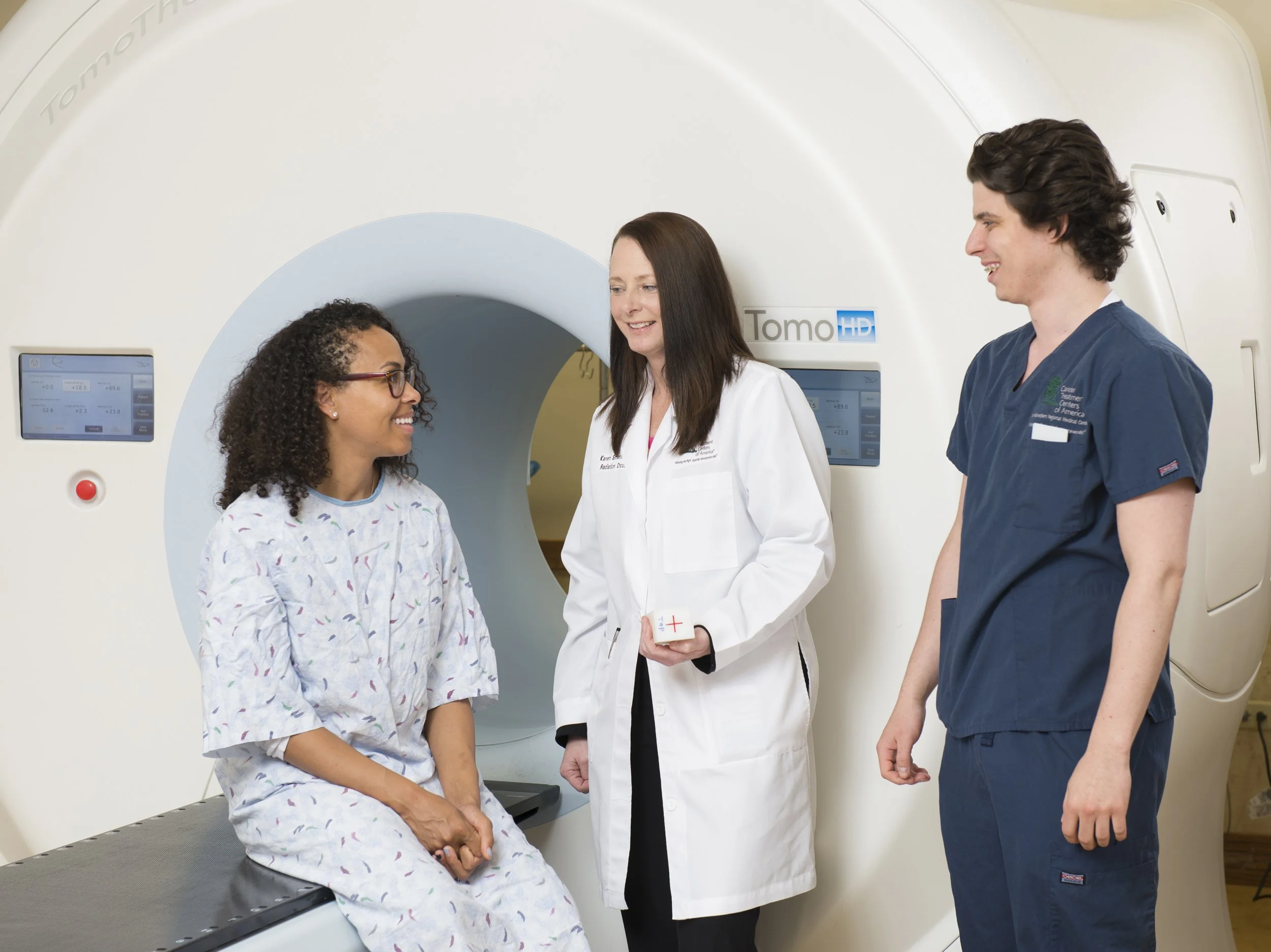 A patient sitting on an examination table talking to two medical professionals in front of a CT scanner.