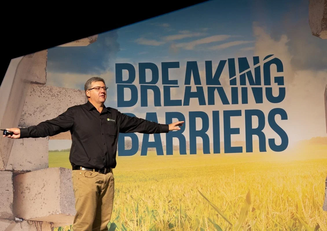 A speaker giving a presentation in front of a large screen displaying the words "Breaking Barriers" against a field and sky background.