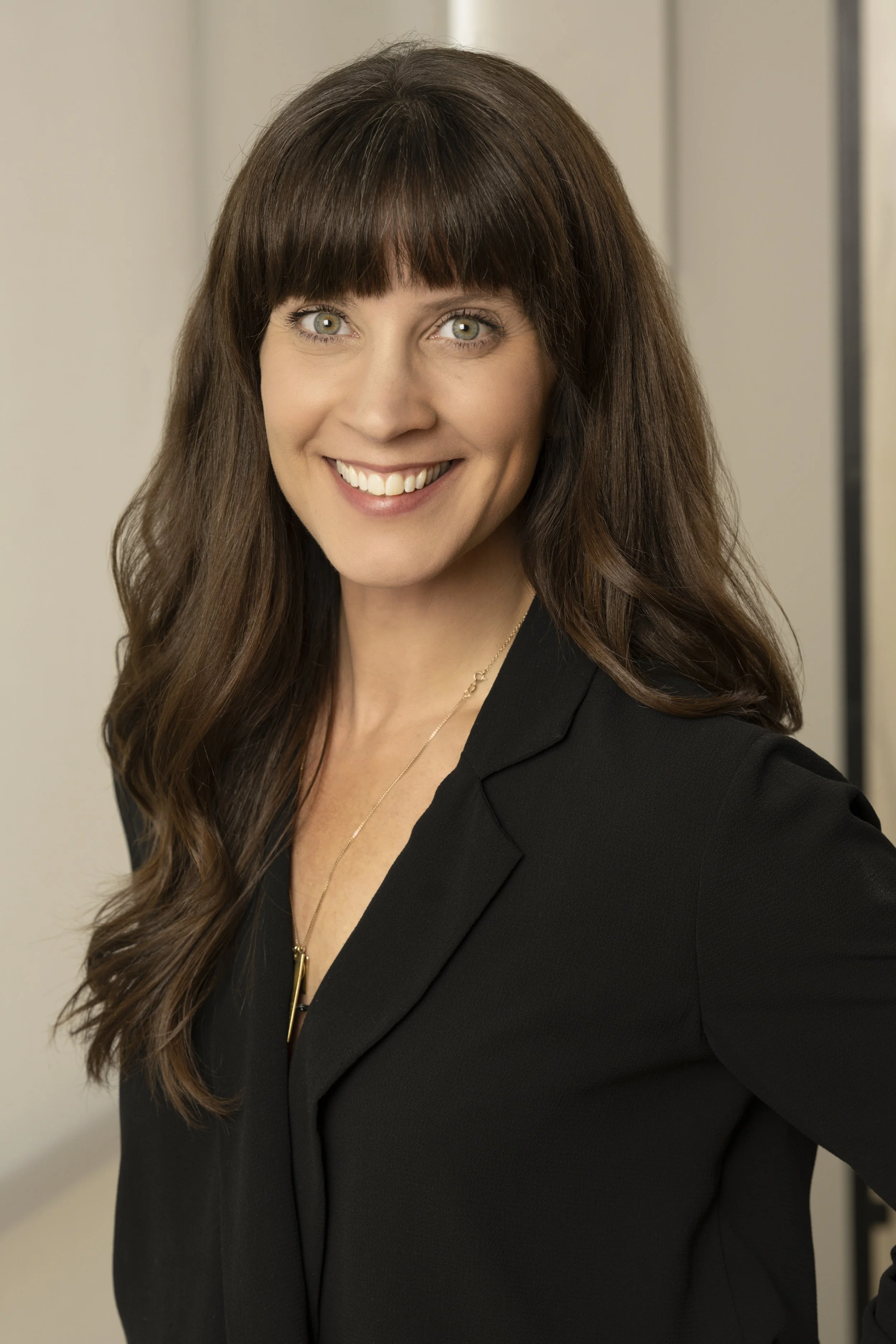 A woman with long brown hair and bangs, wearing a black blazer and a delicate gold necklace, smiling in an indoor setting.