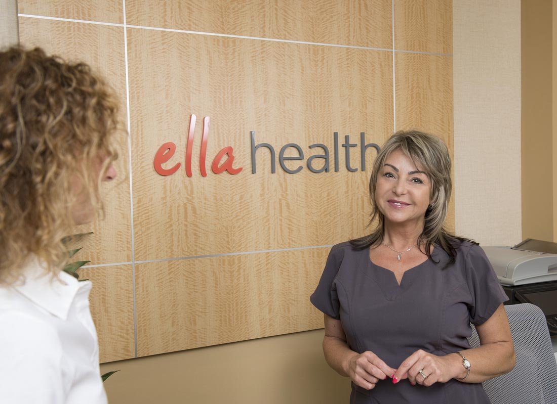 A woman in gray scrubs standing in front of a beige wall with the 'ella health' logo, talking to another woman with curly hair, in an office environment.