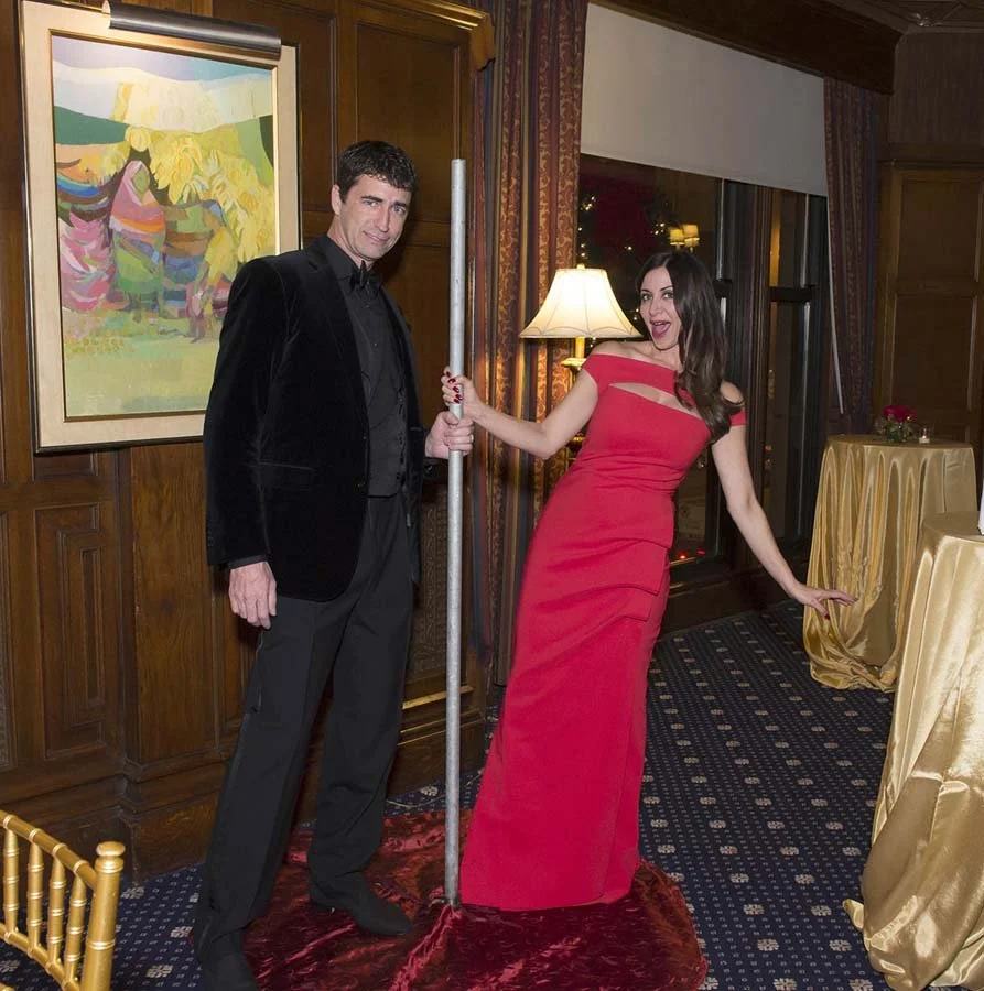 A man and a woman at a formal event holding a pole, with the woman leaning back and smiling. The man is dressed in a black suit, the woman in a red gown. The setting is a decorated room with wood paneled walls, a large colorful painting, a window, a 