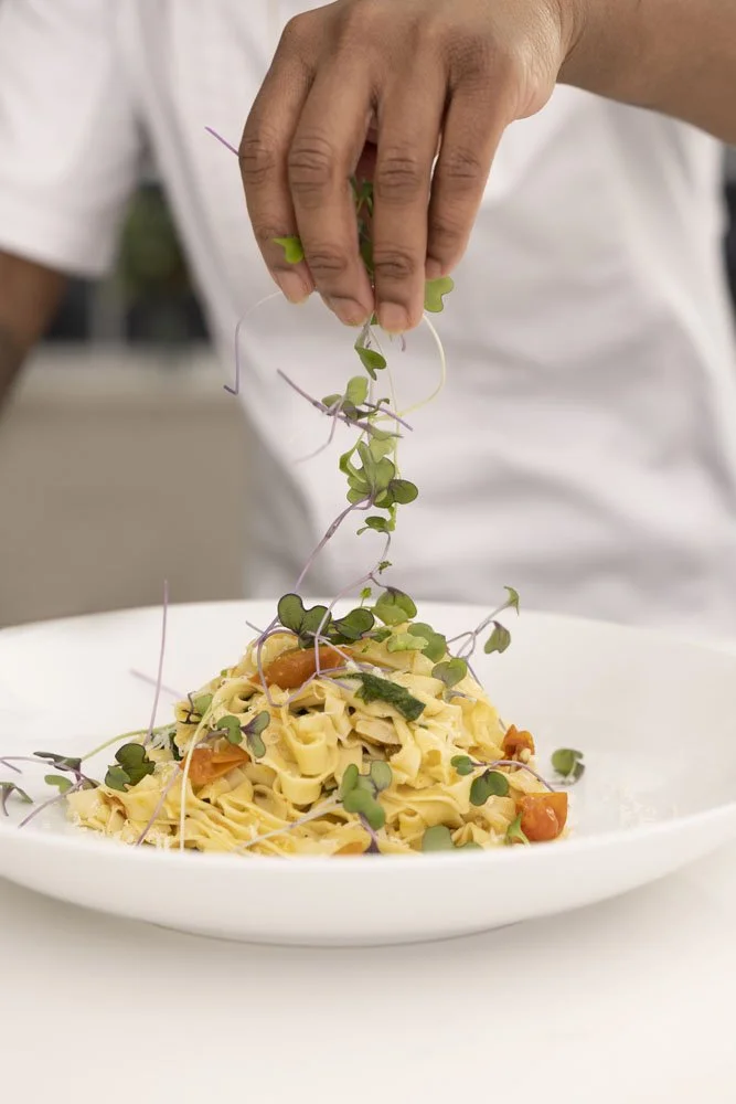 Hand garnishing pasta with microgreens on a white plate for a personal branding photo session.