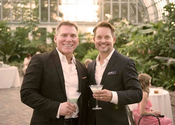Two smiling men in suits holding cocktails at an indoor event with greenery and other attendees in the background.