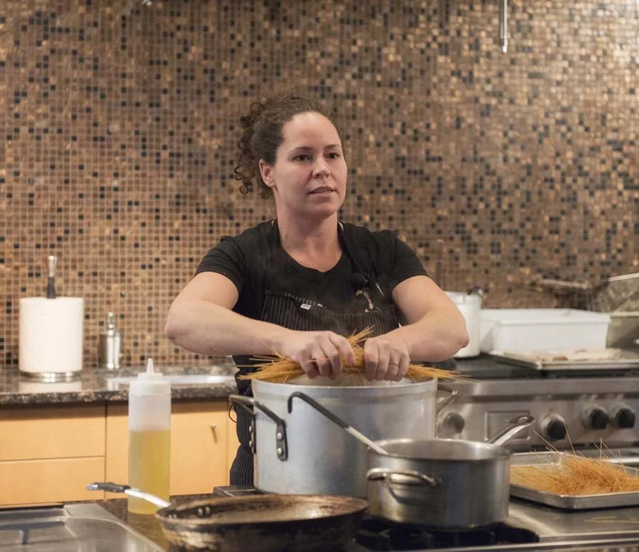 A woman in a black shirt and apron making pasta in a commercial kitchen with a mosaic tile wall for a corporate event.