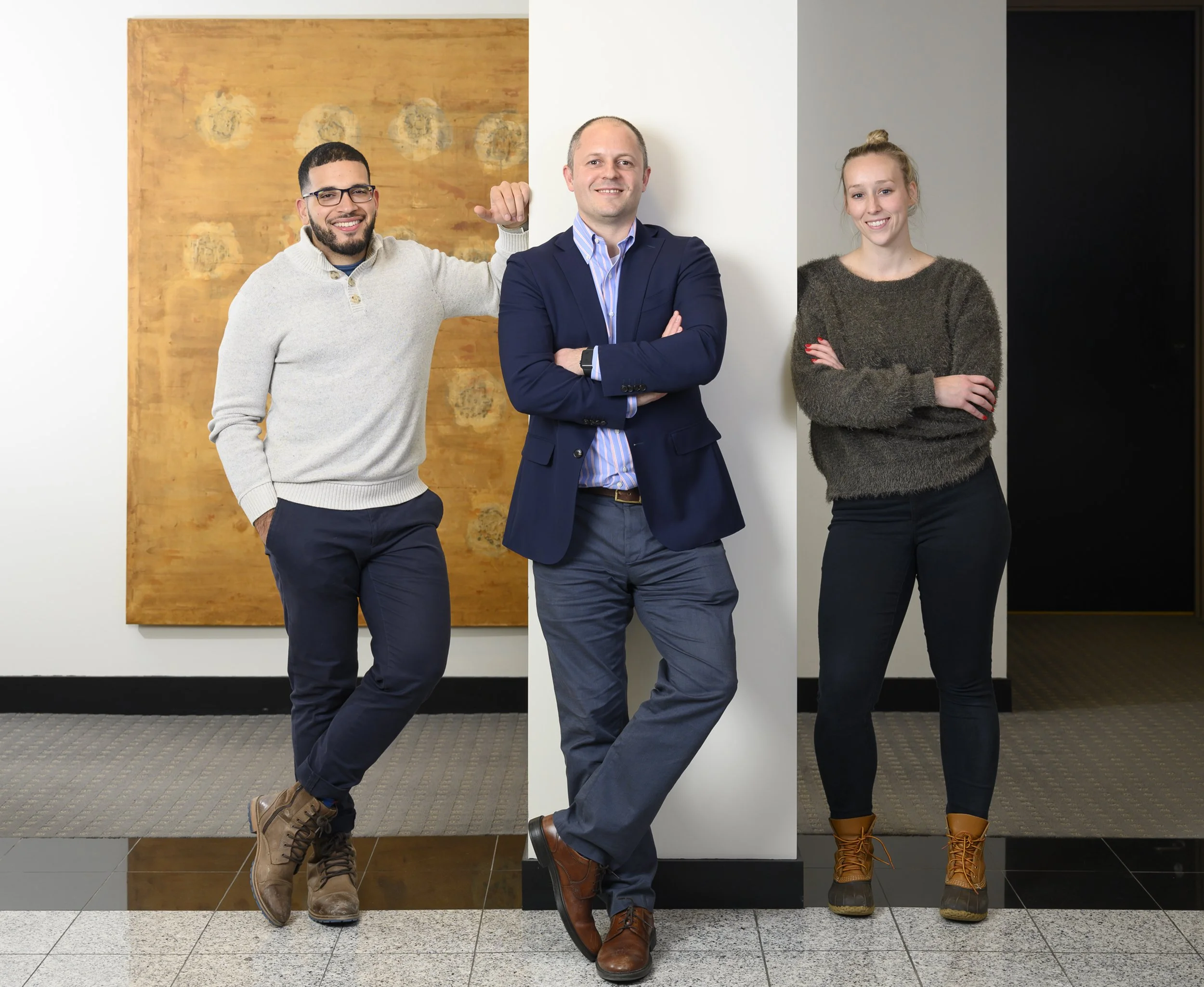 Three people standing together in an indoor setting with artwork and a white column behind them. The man in the middle is wearing a navy suit and smiling with arms crossed, the man on the left has glasses and is dressed casually with a light gray swe