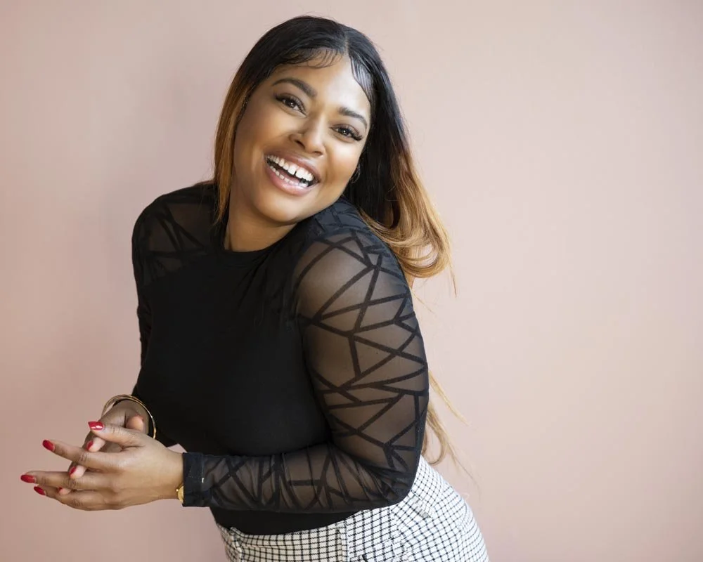 A smiling woman with long, wavy hair, wearing a black top with geometric sheer sleeves and a checkered skirt, standing against a light pink headshot background.