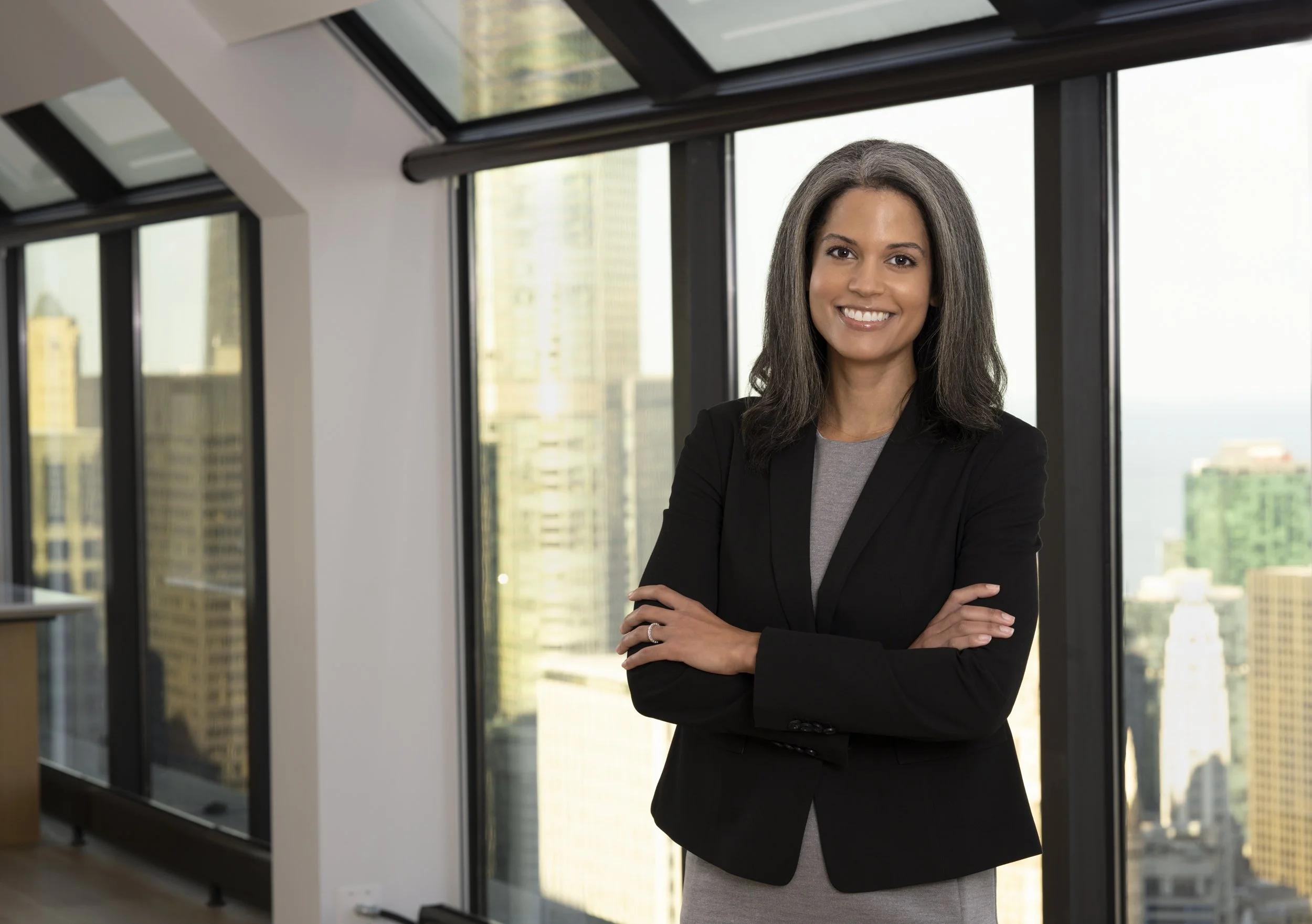 A woman with shoulder-length gray hair smiling with arms crossed in a modern office with large floor-to-ceiling windows showing a city skyline.