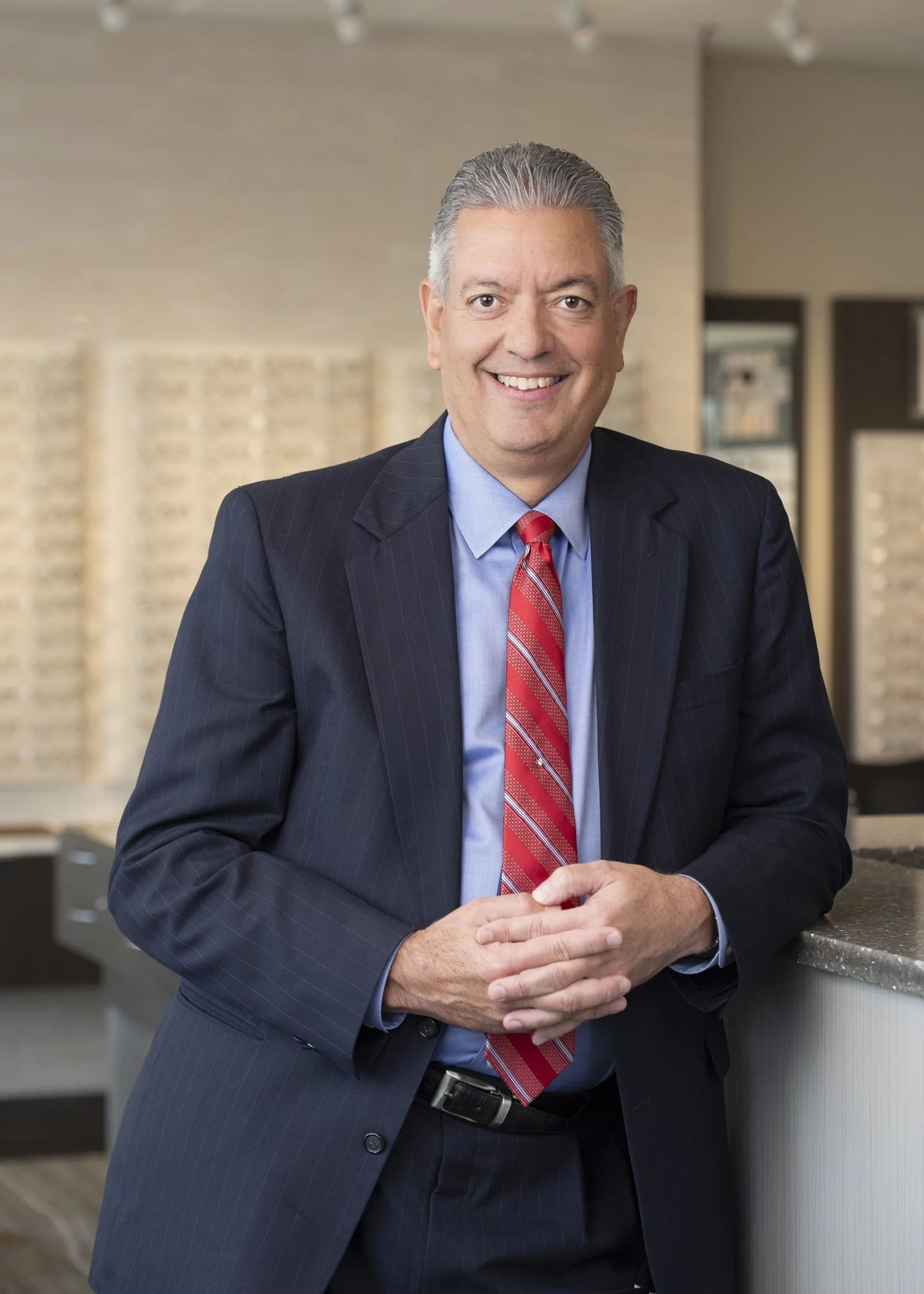 ophthalmologist portrait taken by a Chicago photographer in a dark pinstripe suit and red tie smiling, standing at a reception desk in a modern office setting.