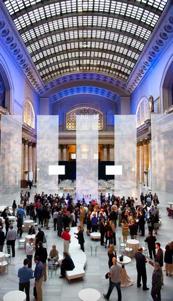 Interior of a large, elegant building with a glass domed ceiling, marble pillars, and a gathering of people at tables.