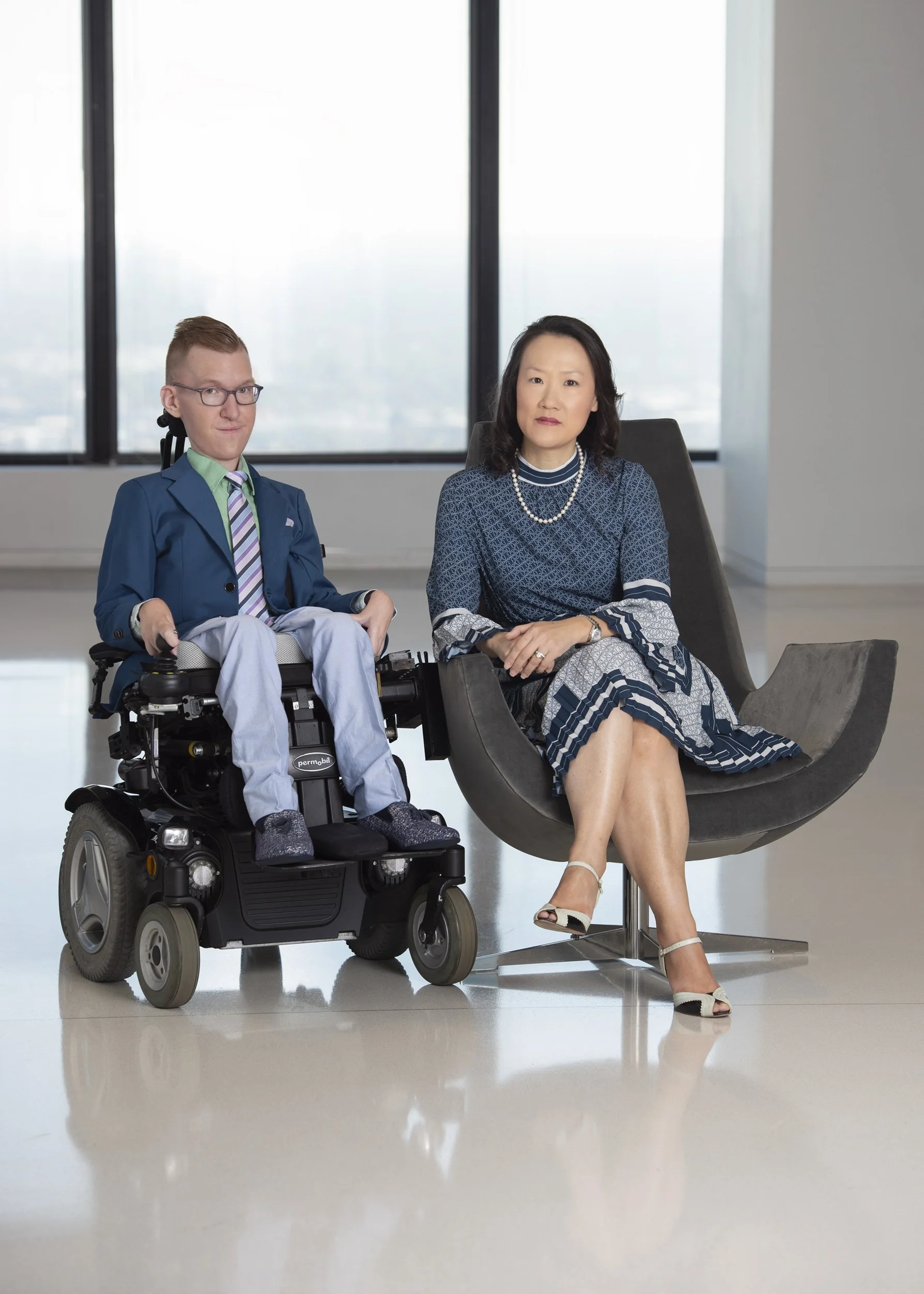Group portrait photography of a woman sitting in a modern office chair next to a man in a motorized wheelchair, both in a bright office with large windows in the background.
