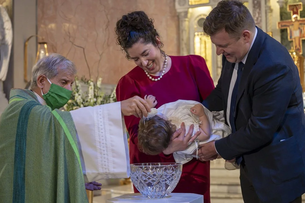 A baby being christened or baptized by a priest during a religious ceremony, with a woman and a man holding the baby, inside a church with ornate decorations and gold accents.