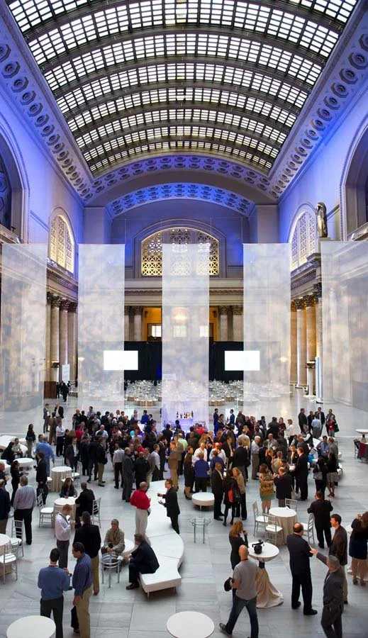 People gathered in a spacious, elegant hall with a high arched glass ceiling, tall marble walls, and large decorative windows, attending a social or corporate event.