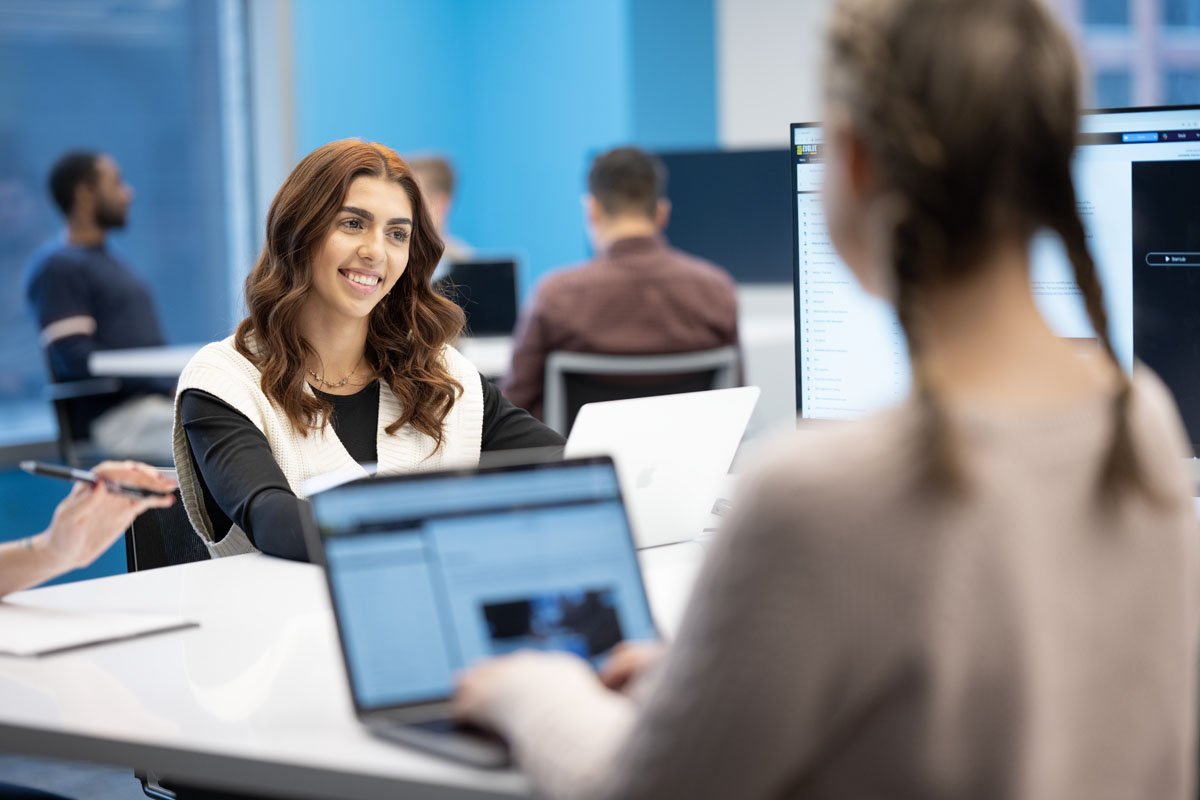 Two women having a conversation at a modern office, with one woman smiling and engaging while the other is partially visible and working on a laptop, with other employees working in the background.