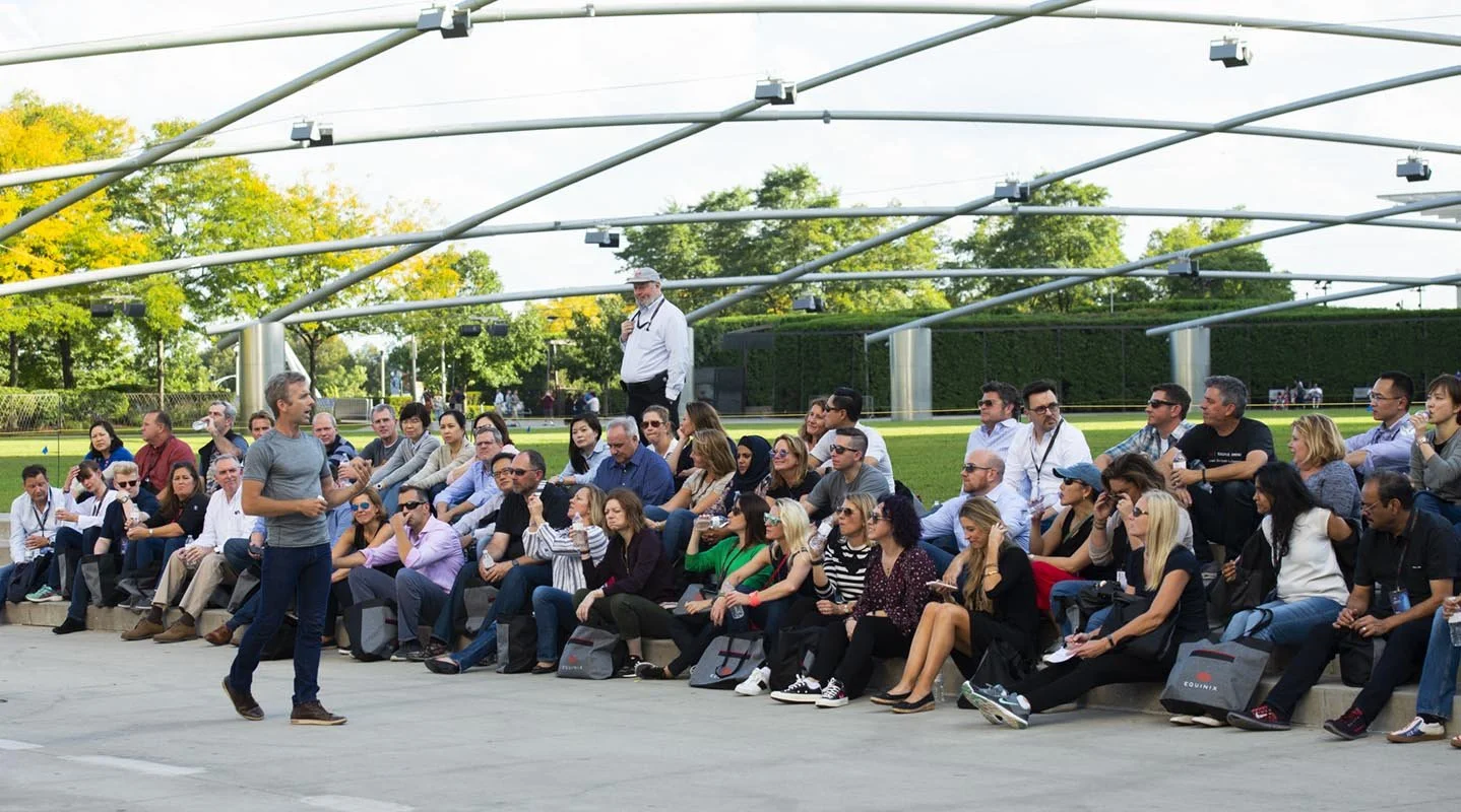 A man in a gray shirt and jeans speaking to an outdoor audience seated on steps, with a speaker in the background and a modern park structure overhead.