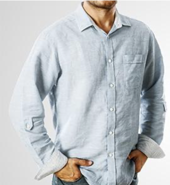 A man wearing a light blue button-up shirt with rolled-up sleeves, standing with hands in pockets.
