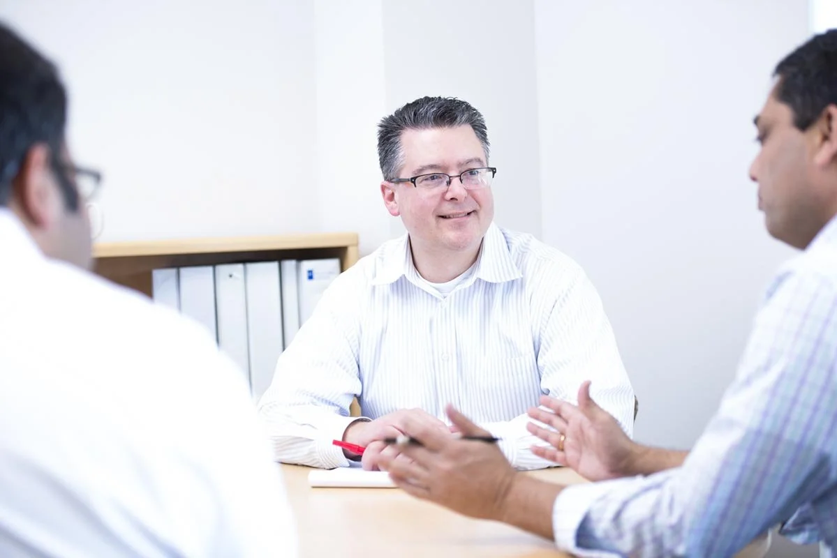 Three men in a meeting room, one in focus with glasses and gray hair, engaging in conversation with two other men, all dressed in business casual attire.