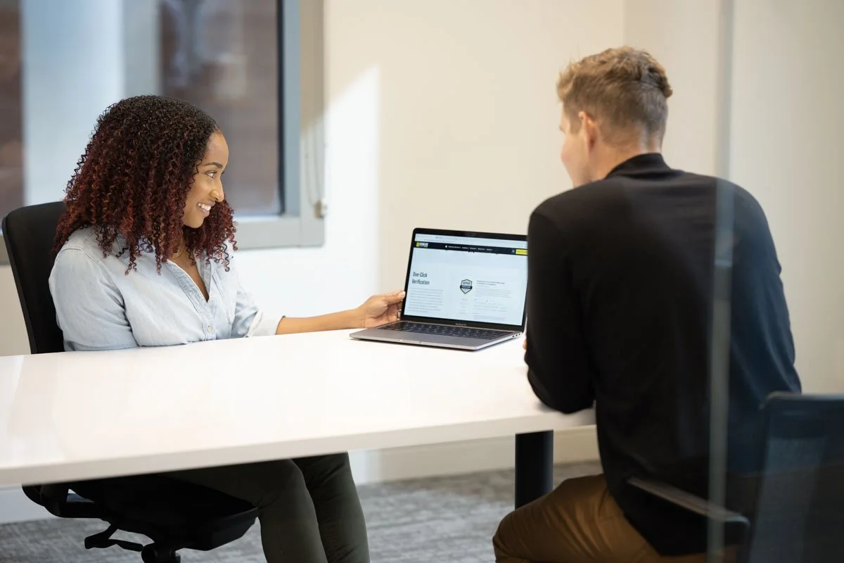 A woman and a man sitting at a white table during a meeting, with the woman showing something on a laptop to the man, in an office setting.