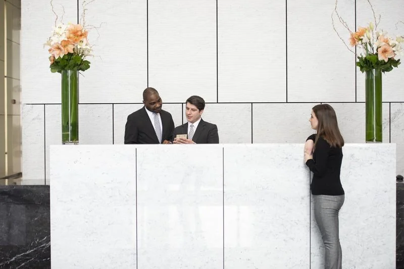 A woman checks in at a hotel reception desk with two men in suits assisting her, decorated with tall floral arrangements on the wall behind.