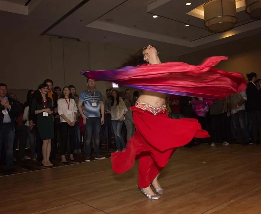 A woman in a red belly dance costume spins and twirls with flowing red and purple scarves in front of an audience at an indoor event.