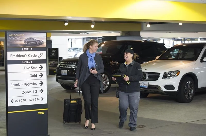 Two women walking in a parking garage, one with luggage, chatting and smiling, near a sign showing directions and Hertz car rental