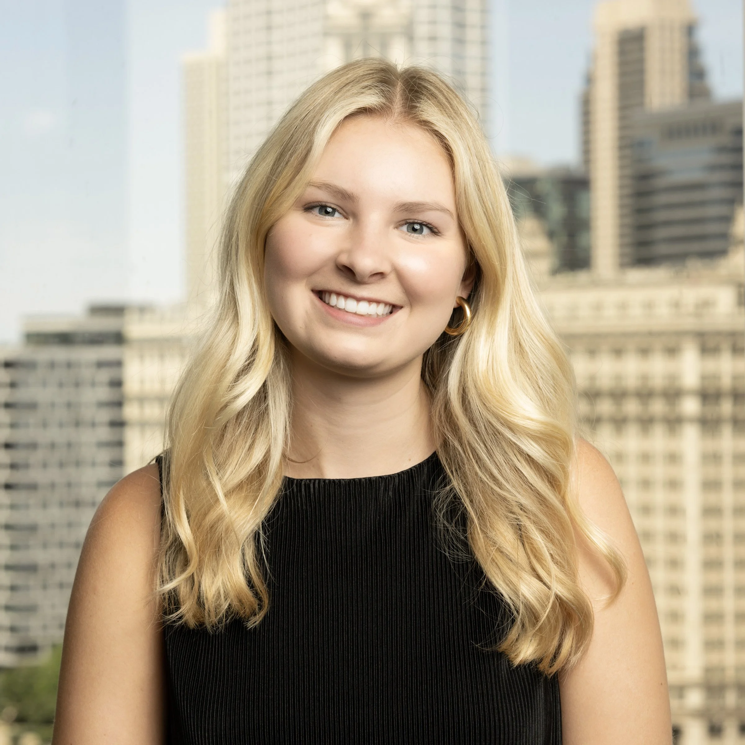 A headshot of a marketing professional woman with long blonde hair, wearing a black sleeveless top, smiling in front of a Chicago cityscape background with tall buildings out of focus.