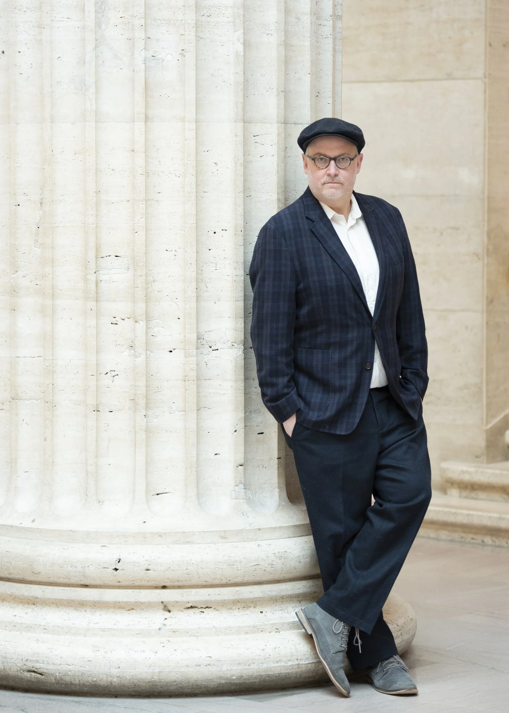 A man with glasses, a black beret, and a gray beard stands with his hands in his pockets against a large stone column, wearing a blue plaid blazer, white shirt, dark pants, and gray shoes.