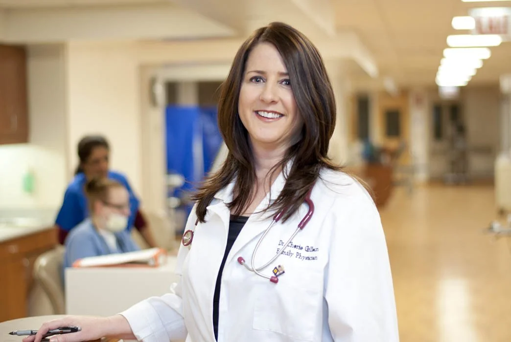 Female doctor in a hospital hallway, smiling at camera, wearing a white coat and stethoscope, with medical staff in background.
