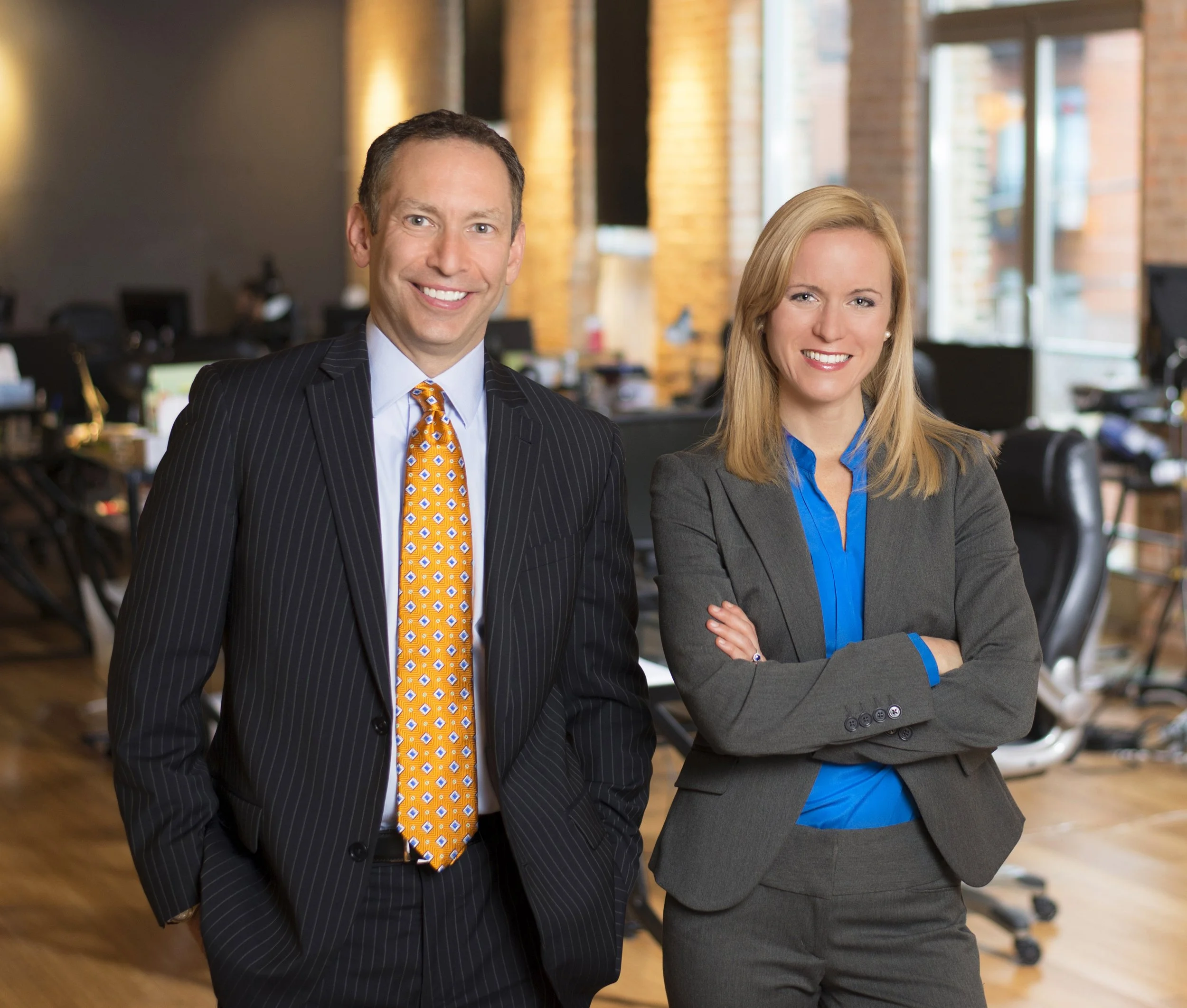 Two professionals, a man and a woman, standing in an office with desks and computers in the background, smiling at the camera.
