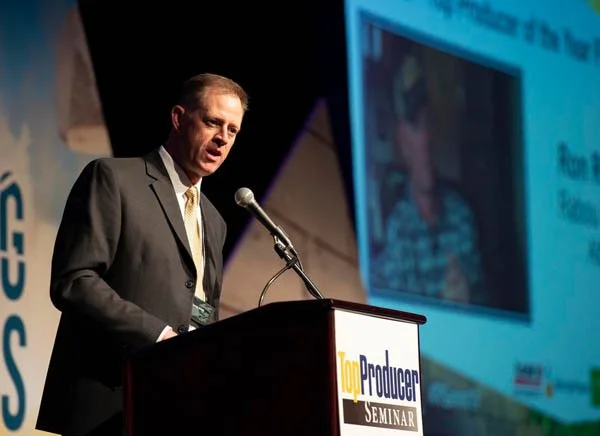A man in a suit speaking at a podium during a seminar or conference with a large screen displaying a presentation in the background.