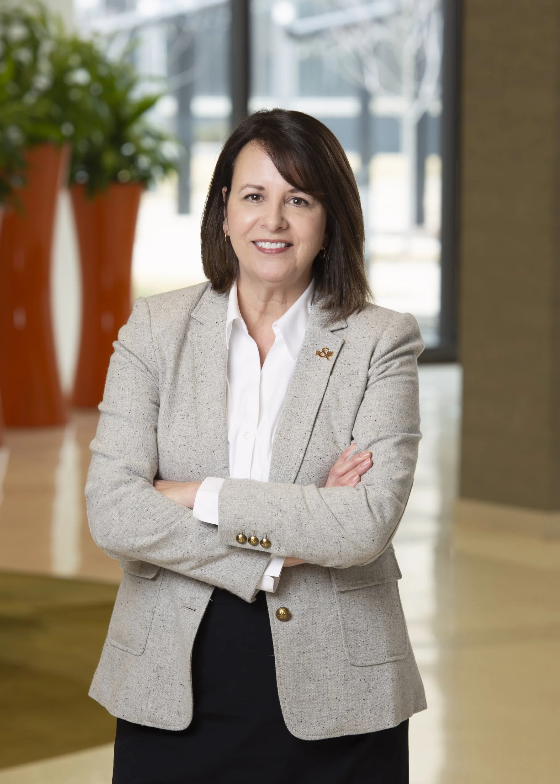 A professional woman with shoulder-length dark brown hair, wearing a light gray blazer with gold buttons, a white blouse, and black pants, standing with arms crossed in a modern office lobby with large windows and decorative orange vases in the backg