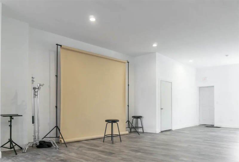 An empty room with a yellow backdrop on a stand, two black stools, and photography equipment against white walls and wooden flooring.