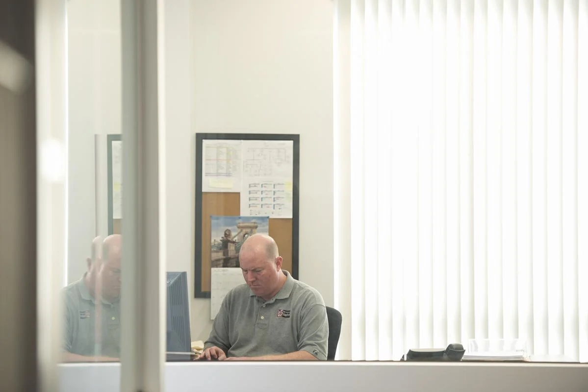Office photos taken for a website of a bald man sitting at an office desk working on a computer, with window blinds behind him and a bulletin board on the wall.