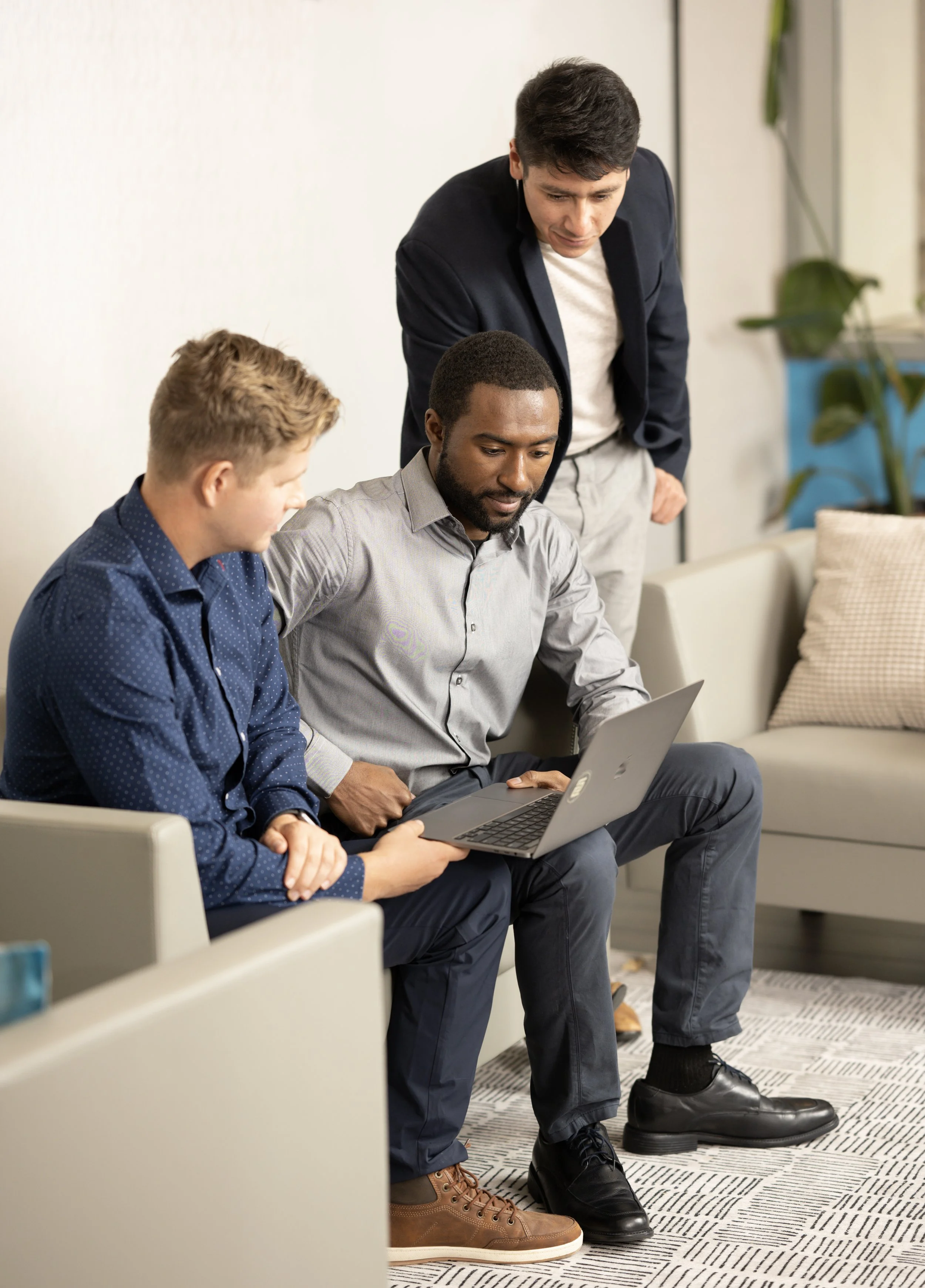 Three men collaborating on a laptop in an office lounge, one seated and two standing, discussing work.