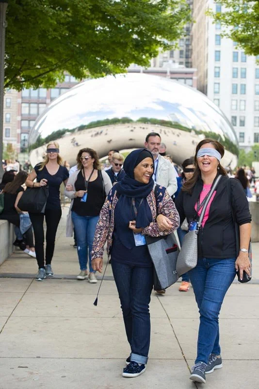 People walking at Millennium Park in Chicago with the Cloud Gate sculpture in the background, some wearing blindfolds during a team building exercise.