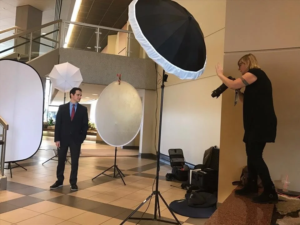 Photo of a professional photo shoot setup with a man in a suit posing, large umbrellas for lighting, and a female photographer taking pictures inside a modern building.